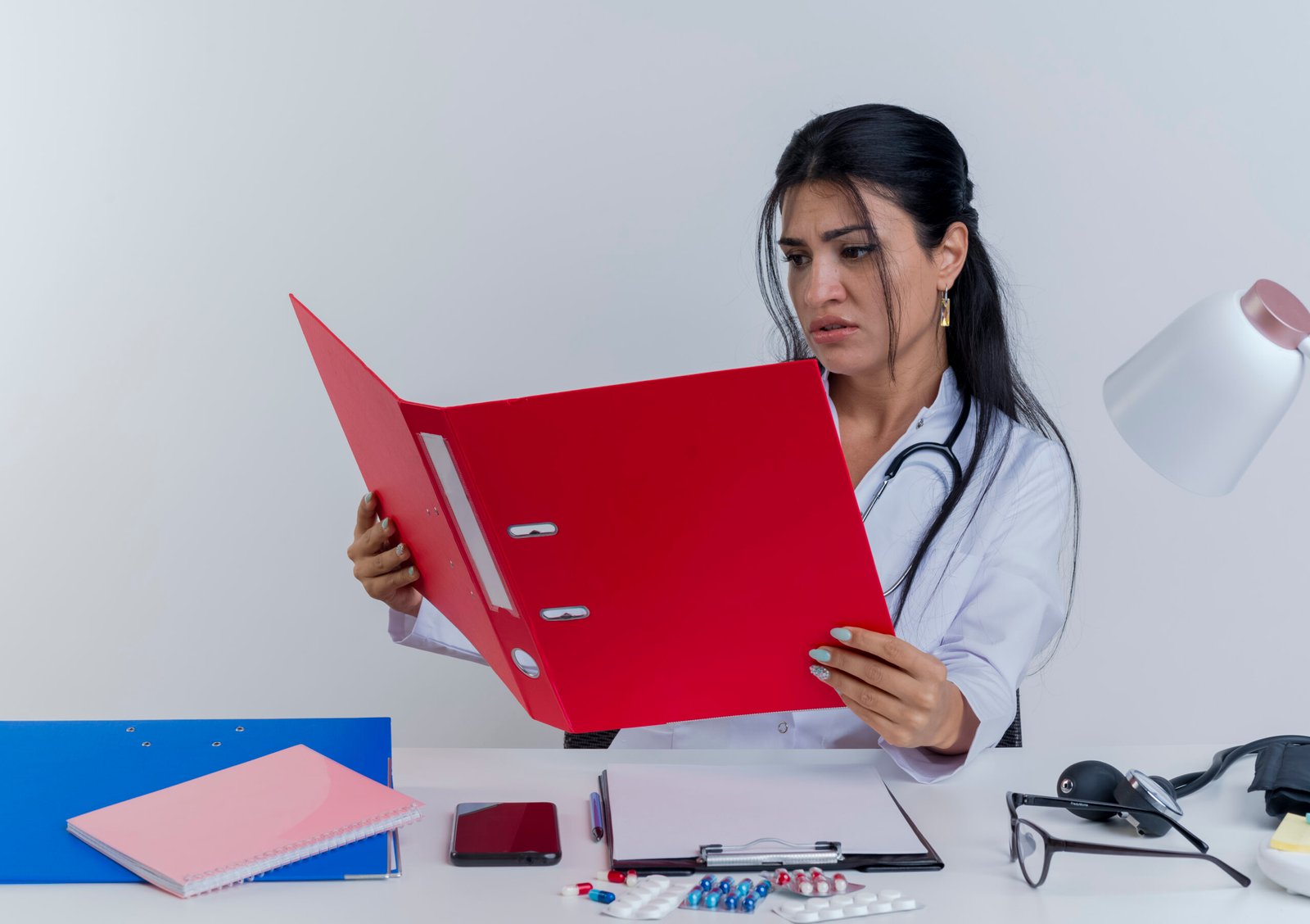 frowning-young-female-doctor-wearing-medical-robe-stethoscope-sitting-desk-with-medical-tools-holding-looking-folder-isolated